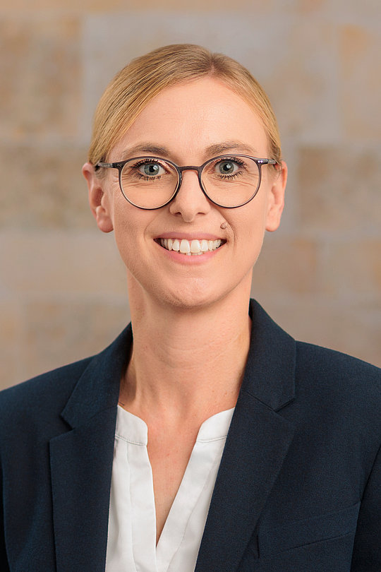 Frau mit Brille und blondem Haar in dunkelblauem Blazer und weißer Bluse lehnt an einer Wand und lächelt