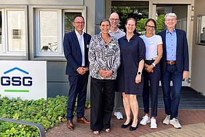 Sechs Personen in Business- und Freizeitkleidung stehen vor einem Gebäude mit einem Schild, auf dem 'GSG' steht